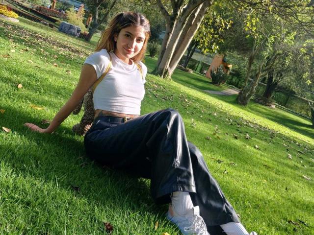 Kaiix is sitting on grass with her ass up, wearing a white top, black pants, and white shoes, looking at the camera with a smile, in a park.