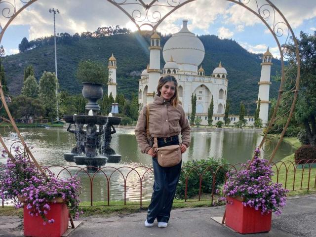 Kaiix stands with a tan jacket, blue jeans, and white sneakers, holding a beige purse, in front of a fountain and a small, white, domed building, surrounded by flowers.