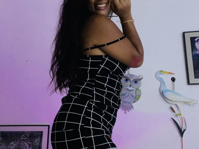 KeyklinOne with dark skin, long black hair, smiling, wearing a black and white checkered dress, showing her shoulder and upper back, her ass slightly visible.