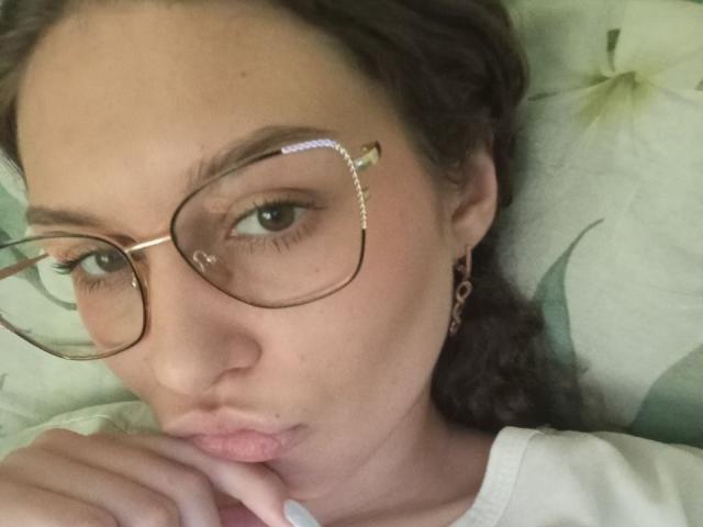 CrisZiebell with glasses, pouty lips, finger near mouth, brown curly hair, light skin, wearing white shirt, earrings, looking at camera, slightly parted lips, subtle makeup.