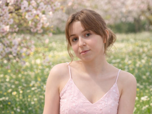 SofiaLuxury with light brown hair, wearing a pink lace spaghetti-strap top, showing small tits and a cute face, standing in a flower-filled meadow.