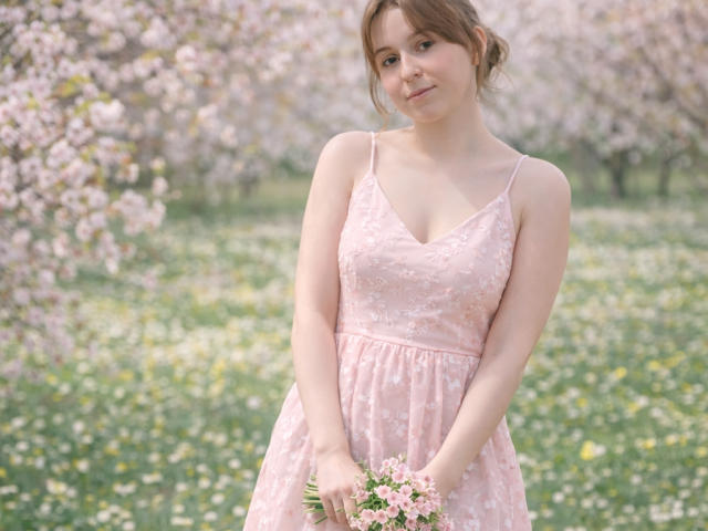 SofiaLuxury with pale skin and brown hair, wearing a pink floral dress, holding pink flowers, standing in a blooming orchard, smiling softly, showing small tits and a cute face.