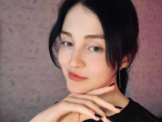 MakedaRiedell has pale skin, black hair, and gray eyes. She's smiling with black nails, a black shirt, and a silver hoop earring, touching her chin.