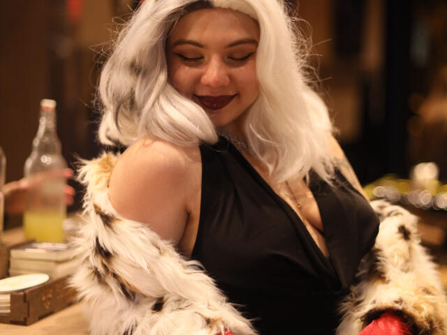 Mettis, con cabello blanco, lápiz labial oscuro, top de tirantes negros y abrigo de piel, sonriendo, sosteniendo una botella, mostrando sus senos, sentada a una mesa con una bebida..