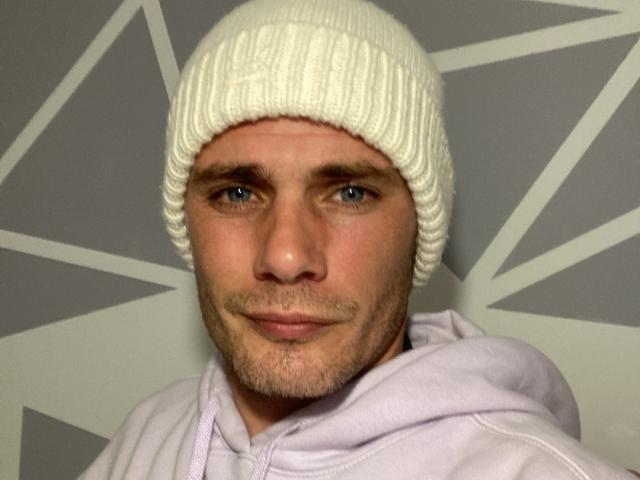RocosiCharly heeft een witte gebreide muts, blauwe ogen, een lichte baardgroei en draagt een lichtpaarse hoodie. Zijn gezicht is enigszins serieus, met een lichte grijns..