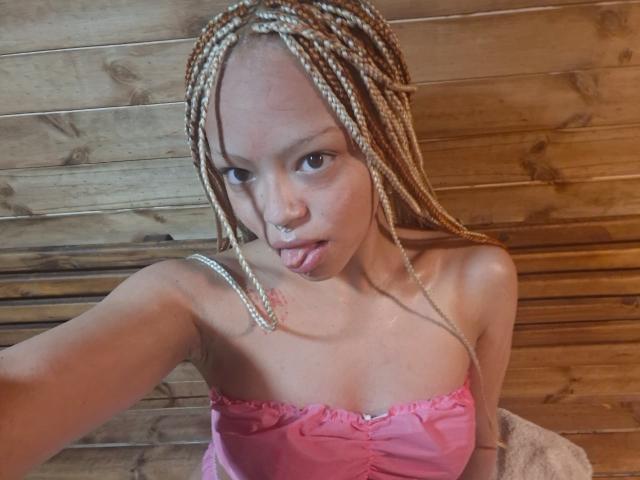 KeiryGinger with blonde braids, pouting lips, pink strapless top, showing small tits, and a cute nose ring, in a wooden room, taking a selfie.