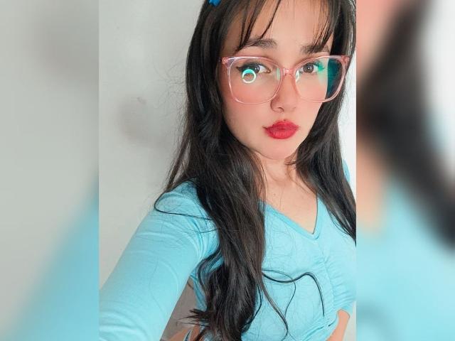 NataliaRivas with long black hair, pink glasses, red lipstick, and blue top, showing her tits in the mirror selfie. Her reflection is slightly blurred on the right.