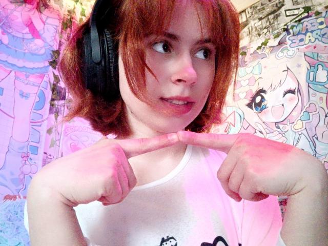 MireilleSpotwood with red hair, wearing headphones, pointing at her chin with both index fingers, wearing a white shirt, looking to the side, in a brightly lit room.
