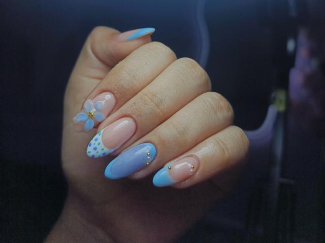 LissyCarter's hand with pastel blue and pink nails, featuring a small flower and gold studs on the ring and pinky, and blue polka dots on the middle nail.