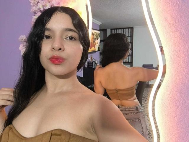 LissyCarter with long black hair, red lipstick, tan strapless top, mirror reflecting her bare back, small tits, standing in purple room with wavy mirror light.