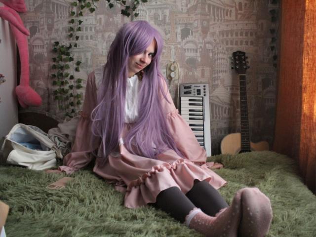 MireilleSpotwood with long purple hair, pink dress, and black socks sits on green shag carpet, legs crossed, looking down, in a room with a keyboard and guitar.