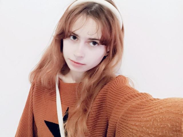 MireilleSpotwood with long red hair, white headband, and orange ribbed sweater, looking slightly annoyed, pale skin, small tits, subtle nose, and soft brown eyes.