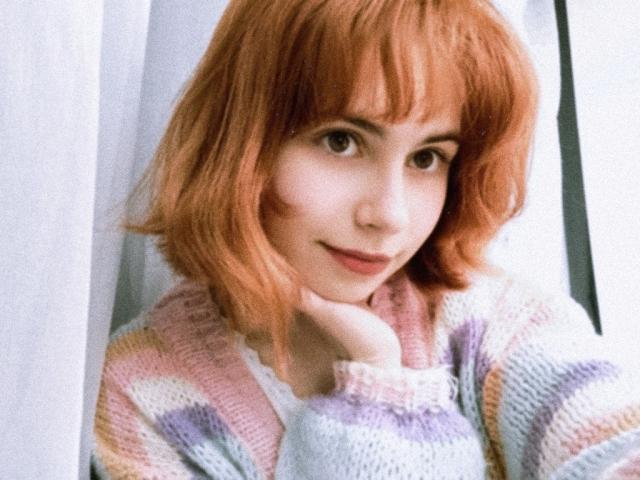 MireilleSpotwood with red hair, wearing a pastel striped sweater, resting her chin on her hand, smiling softly. Her tits are small and her skin is pale.