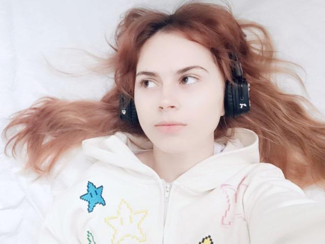MireilleSpotwood with red hair, wearing black headphones, white hoodie with colorful star patches, lying down, looking to the side, pale skin, slight pout, soft expression.