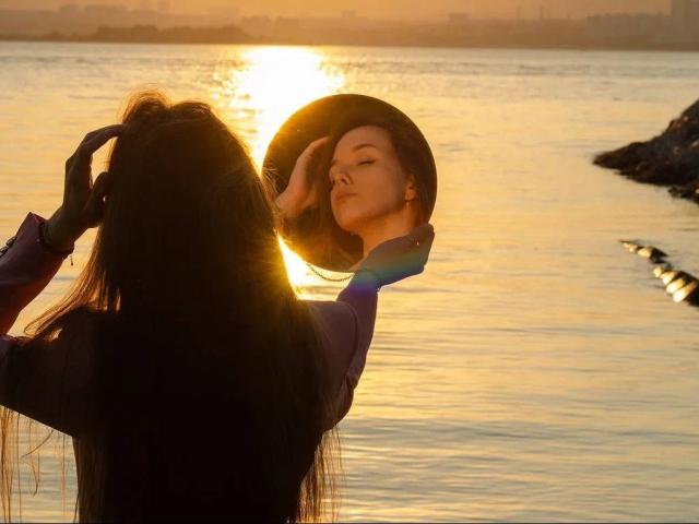 ShaunnaRoepke with long dark hair, wearing a hat, touching it with her right hand, sunlight reflecting off the water, golden hour, serene, elegant, feminine, peaceful.