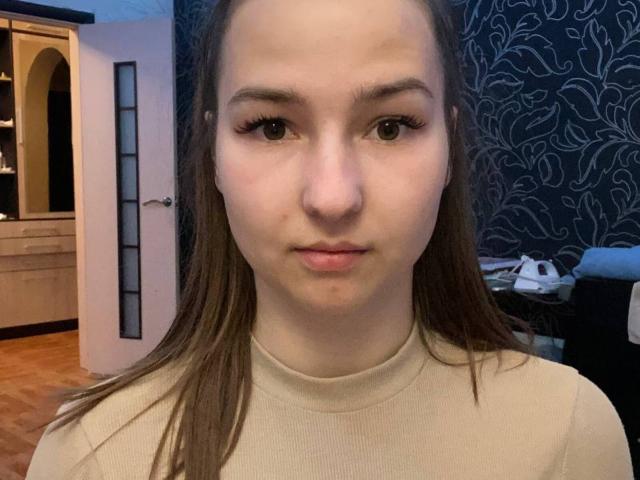Jeanelle Mulholland tiene la piel clara, ojos marrones y cabello largo y castaño. Lleva un suéter de cuello alto color beige, tiene una expresión neutra y un pequeño grano en el labio inferior..