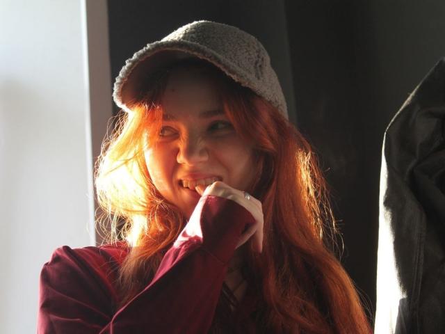 KatiRokisky with red hair, wearing a grey flat cap and red shirt, has sunlight in her face, smiling with her finger near her mouth, looking cute.
