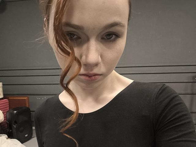 KatiRokisky has pale skin, brown eyes, and a serious expression with a wavy red ponytail, wearing a black shirt, showing her bare neck and small, slightly parted lips.