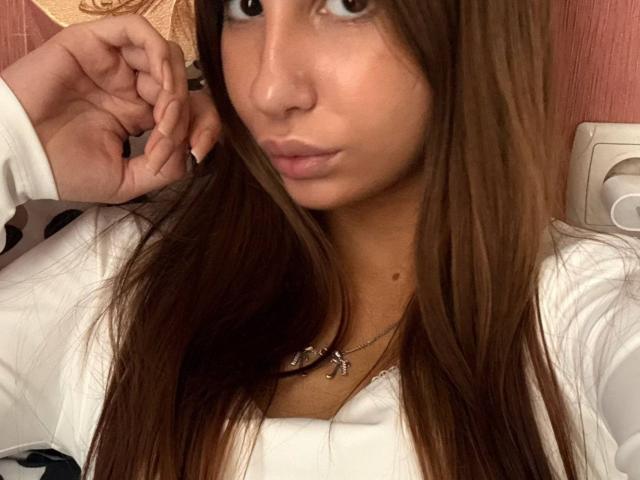 KatherinaEdnie with long brown hair, wearing a white shirt, holding her fingers near her face, looking slightly surprised. She has a necklace with small charms.