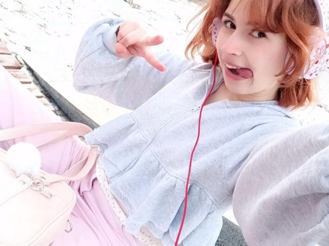 MireilleSpotwood with red hair, sticking out tongue, wearing pink headphones, light blue shirt, pink pants, and pink bag with fluffy pom-pom, making a rock gesture with her finger.
