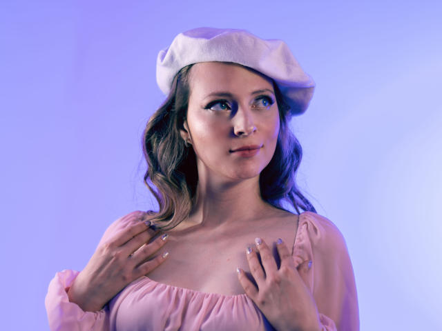 AmbeerWood with wavy brown hair, wearing a pink dress and white beret, hands on chest, blue-purple lit face, looking up, subtle smile, clear nails, soft light.