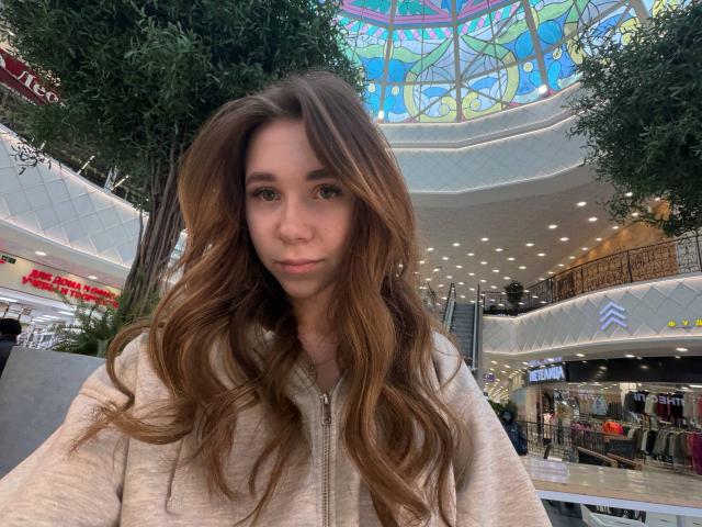 PokBingman has long brown wavy hair, wearing a beige zip-up hoodie, with a neutral expression, standing in a brightly lit mall, featuring a colorful stained glass ceiling.