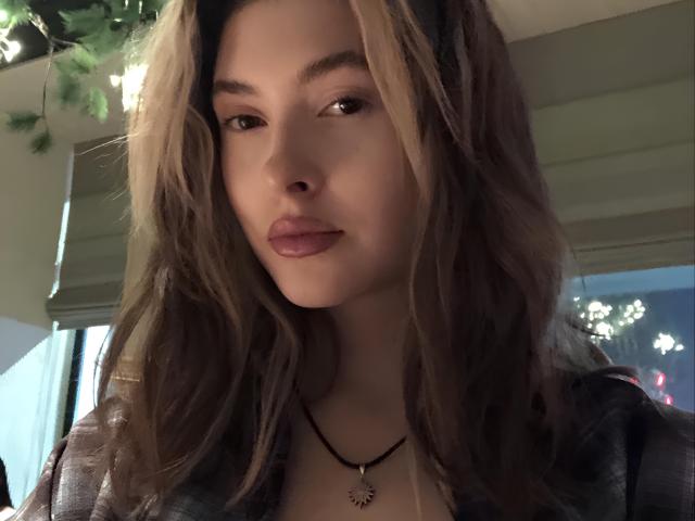 MaryamRisby with wavy brown hair, full lips, and a necklace, wearing a dark shirt, looking into the camera with a subtle, confident expression.
