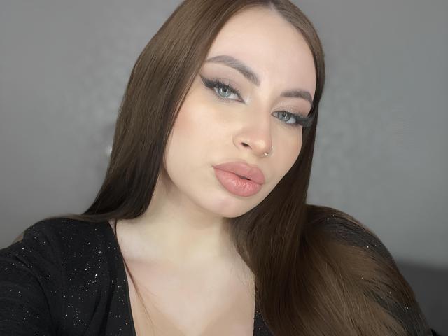 BillyeShambo has pale skin, long brown hair, blue eyes, winged eyeliner, pink glossy lips, nose ring, black sparkly top, looking slightly to the side with a pouty expression.