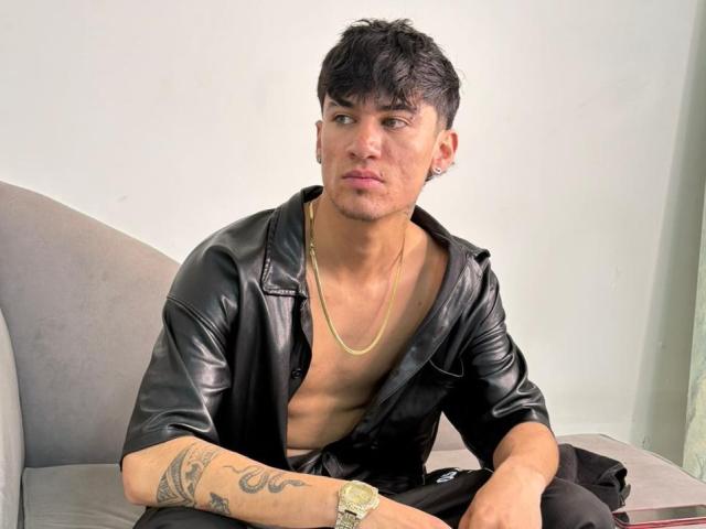 TheBossCrown ist ein attraktiver Mann mit kurzen, schwarzen Haaren, der ein schwarzes Lederhemd, eine goldene Kette und schwarze Hosen trägt. Er hat Tattoos am Arm und eine Uhr..