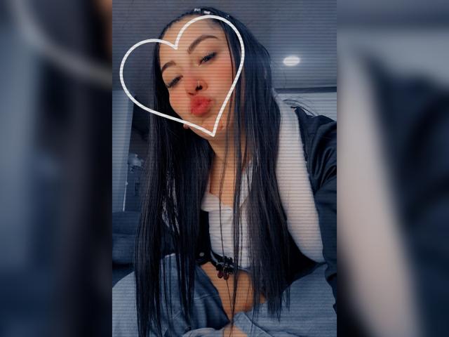 Alexa Adams, con cabello negro largo, labios carnosos y un emoji de corazón sobre su rostro, vestida con una blusa blanco y negro, sentada con las manos frente a ella, fondo borroso..