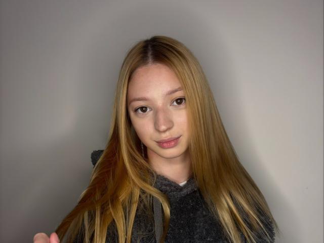 Pantelina mit langen, blonden Haaren, die einen dunkelgrauen Hoodie trägt und ihr Haar mit einem sanften Lächeln zurückhält, helle Haut, braune Augen und ein süßes, unschuldiges Aussehen..