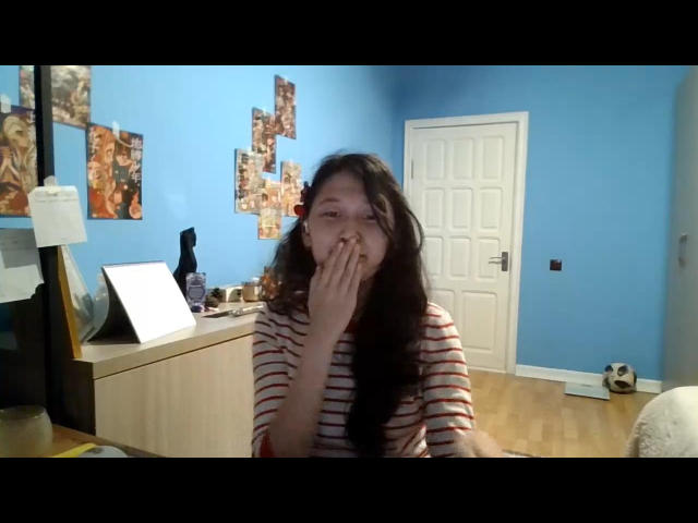 LennaScripter, met lang donker haar, draagt een rood-wit gestreepte blouse en bedekt haar neus en mond met haar hand, ze kijkt lichtelijk geïrriteerd. Haar borsten zijn klein en ze is schattig..
