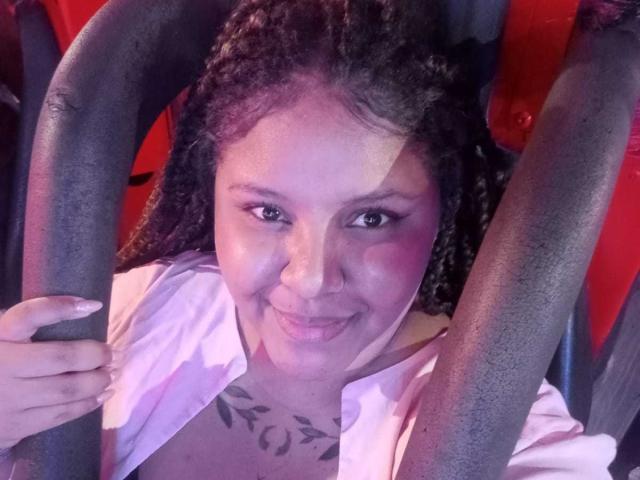 AkiraHeat with dark curly hair, pink shirt, and tattoo on chest, smiling with tongue out, holding gray metal bars, purple and pink light on face, pink lipstick.