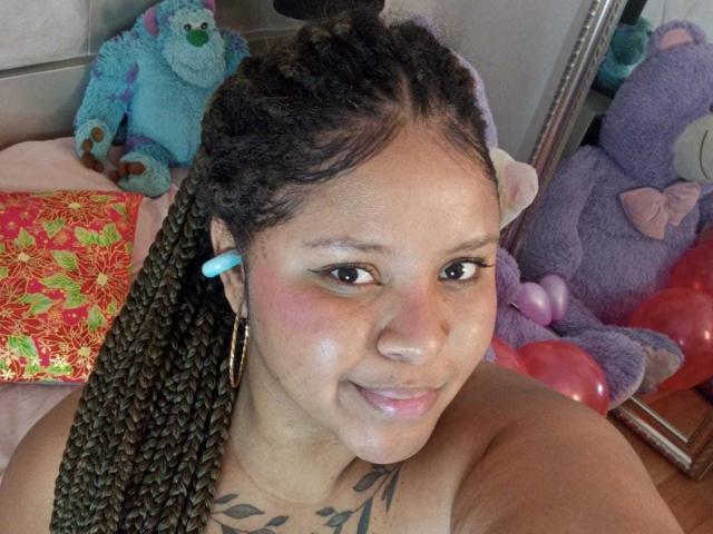 AkiraHeat, con cabello castaño trenzado, audífonos azules, aros grandes, rubor rosa, sonriendo, con un tatuaje visible en el pecho, pezones rosas, sin ropa, rodeada de peluches..