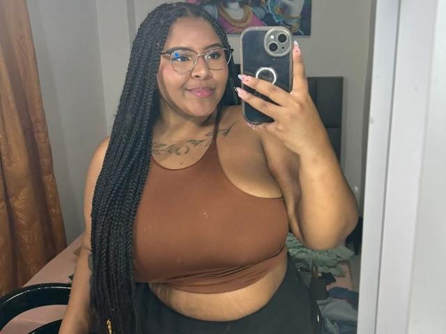 AkiraHeat with long black braids, glasses, brown crop top, showing big tits and a small belly, taking a mirror selfie with a smile, holding a black phone.