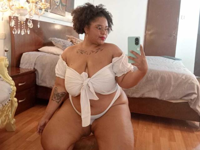 AkiraHeat with curly hair, glasses, white off-shoulder top tied at chest, white thong, tattoo on right arm, holding phone, kneeling, showing large tits and ass, bedroom.