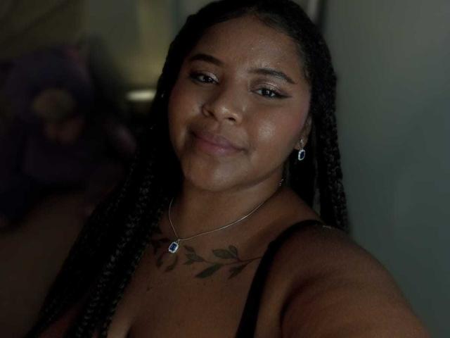AkiraHeat, a dark-skinned woman with long braids, subtle smile, small earrings, necklace, and a leafy chest tattoo, takes a selfie, showing her bare shoulders and black top.