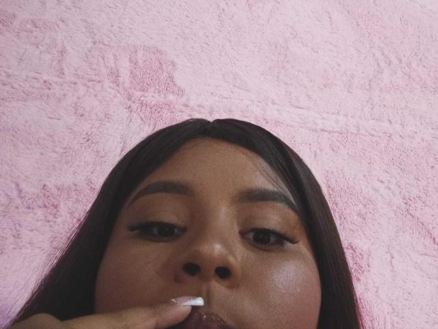 AkiraHeat's face with dark brown skin, long black hair, and thick eyebrows, sucking on a finger with white nail polish, pink textured background, slightly flushed cheeks.