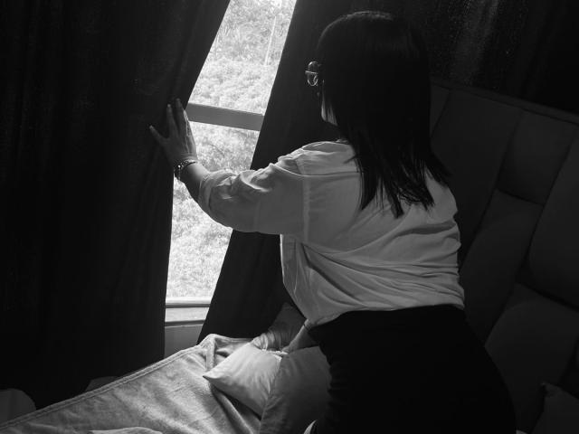 DiannaRosse with glasses, white shirt, black skirt, pulling dark curtain to the side, exposing window, sitting on bed with pillow, in black-and-white photo.