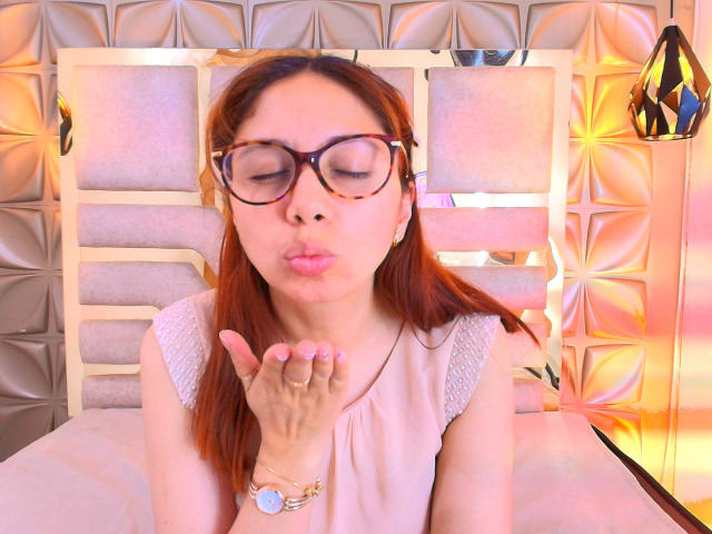 StephanieSun with pink lips puckered, wearing glasses, white sleeveless top, and a pink wristwatch, hands near face, brown hair, gold earrings, warm light.