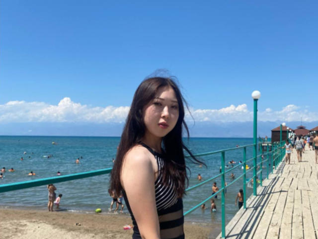 RemediosServille, con lunghi capelli neri, indossa una maglietta a righe nere e bianche e si trova su una passerella sulla spiaggia, guardando verso la fotocamera, con un oceano blu-verde e persone sullo sfondo..