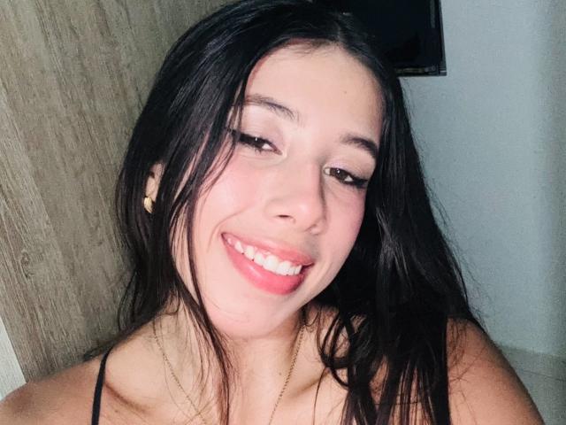 LanaKingsley with long black hair, light skin, and a cute smile, showing pink lipstick and subtle eye makeup, wearing a black strap top and a gold necklace.