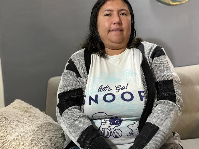 BiancaValli, uma mulher corpulenta com seios de tamanho médio, cabelo preto na altura dos ombros e um sorriso, veste um cardigã listrado sobre uma camiseta com a frase "Let's Go! NOOOOR", sentada em um sofá..