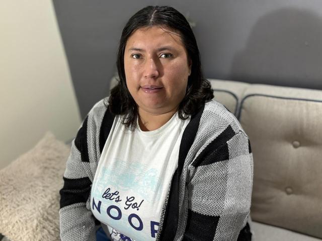 BiancaValli, uma mulher corpulenta com cabelo castanho escuro, vestindo uma camisa branca e um cardigã listrado preto e cinza, sentada em um sofá bege com um travesseiro fofo..
