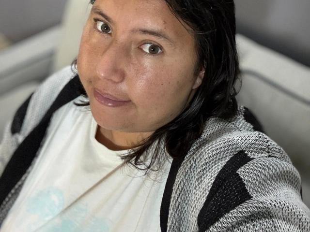 BiancaValli, con la pelle di colore marrone chiaro, i capelli scuri e ondulati, indossa una camicia bianca e un cardigan grigio-nero con motivi, sorride leggermente, ha gli occhi marroni, un trucco leggero e un'espressione serena..