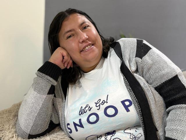 Bianca Valli, eine etwas mollige Frau mit dunklem Haar, trägt einen grauen und schwarzen Strickcardigan über einem weißen Hemd mit der Aufschrift "let's go SNOOP" und lächelt, während sie ihren Kopf anlehnt..