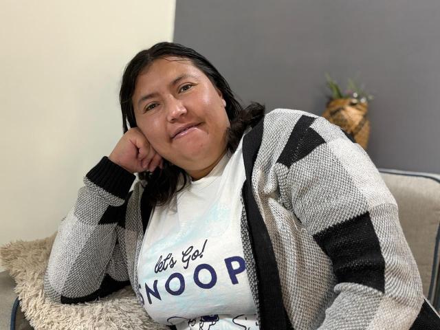 BiancaValli, de complexión robusta, con senos medianos, cabello negro, vestida con un cárdigan gris a rayas y una camisa blanca, sonriendo, con la cabeza apoyada en la mano, sentada en una silla beige esponjosa..