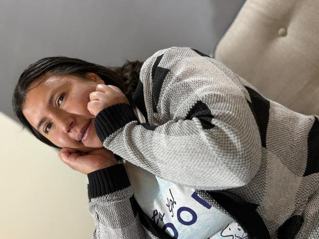 BiancaValli, con cabello oscuro, vistiendo un suéter con estampado gris y negro, con la cabeza apoyada en la mano, sonriendo ligeramente, sentada en una silla beige..