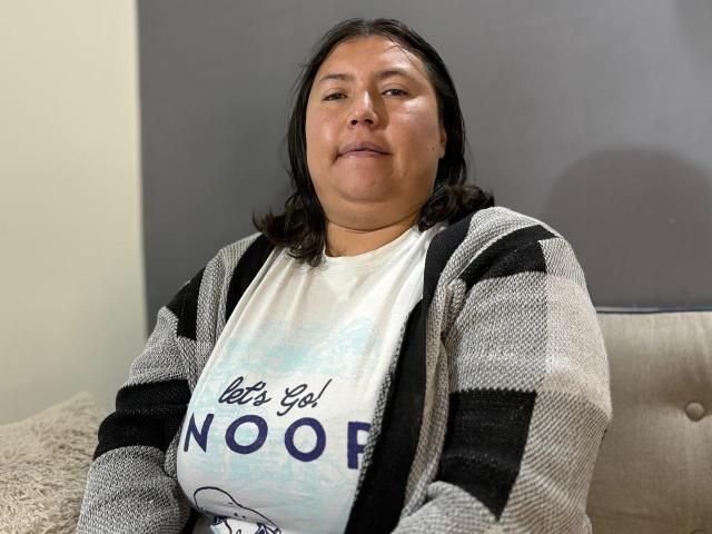 BiancaValli a un visage rond, des cheveux noirs et lisses, et un léger sourire. Elle porte un cardigan rayé gris et noir par-dessus un t-shirt blanc avec l'inscription "let's go NOO!"..