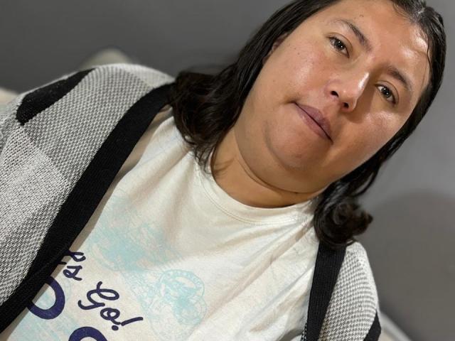 Bianca Valli mit schulterlangem, schwarzem Haar, hellbrauner Haut, einem leichten Lächeln, trägt ein weißes Hemd und eine graue Netzjacke mit schwarzen Trägern auf den Schultern, in einer lässigen Pose, leicht geneigter Kopf..